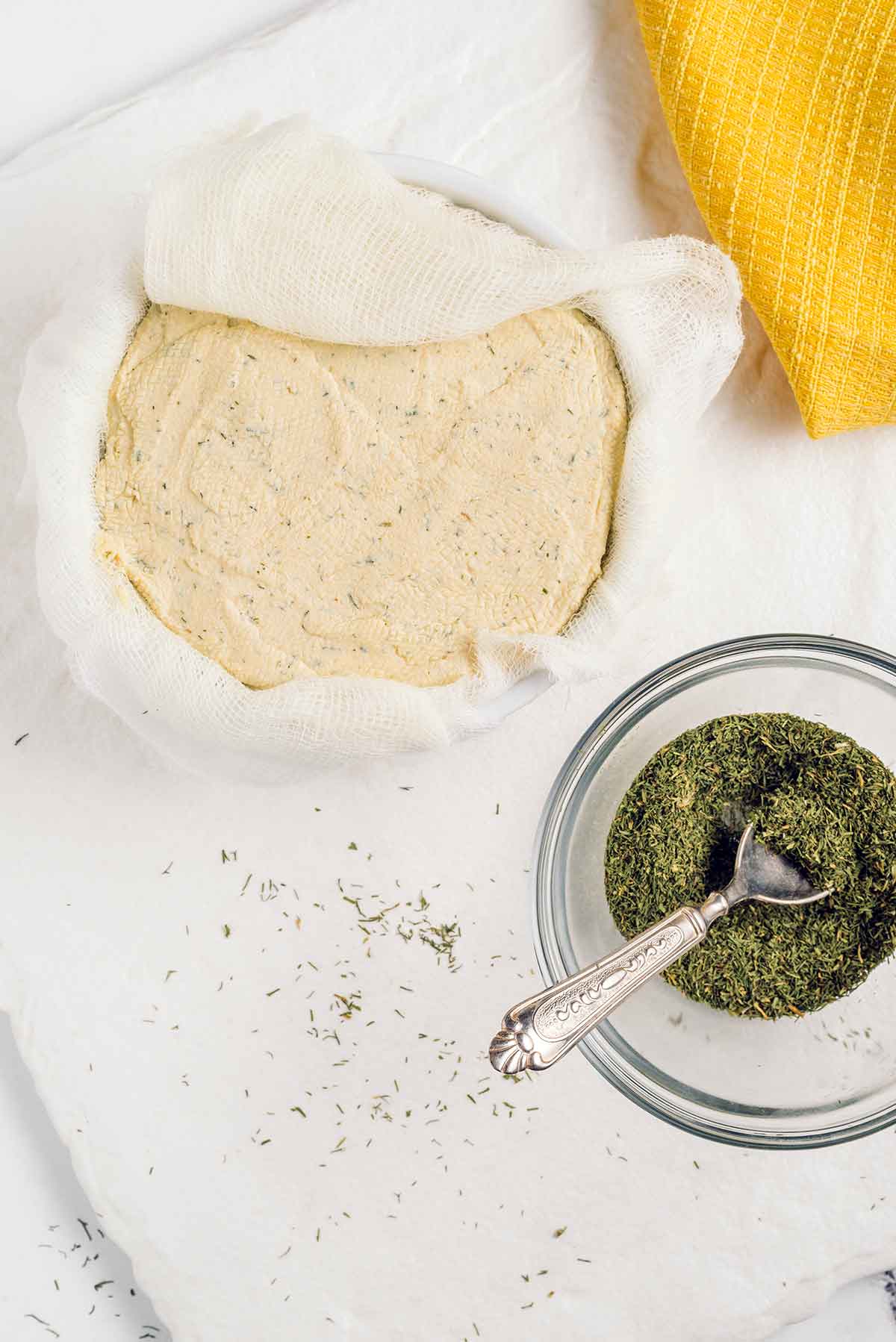 Top down view of vegan cheese wrapped in cheesecloth and resting in a ramekin. Dried dill for garnishing the vegan cheese log fills a small bowl nearby.