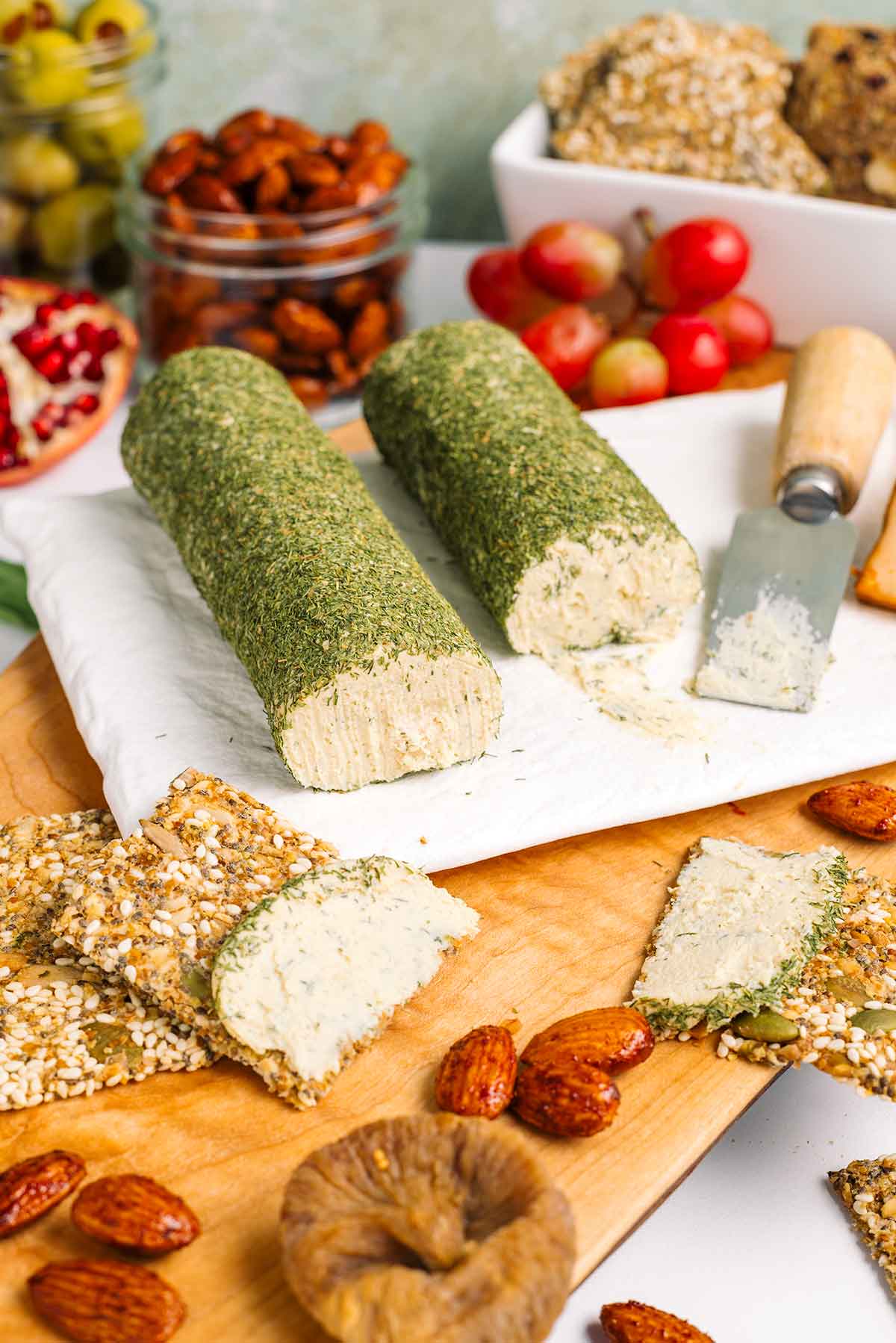 Close up of two vegan cheese logs coated with dried dill. The cheese is white, soft, and spreadable. Crackers spread with cheese rest on a charcuterie board of fruits, nuts, smoked tofu, and olives.