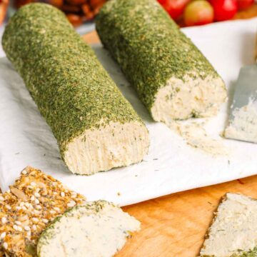 Close up of two vegan cheese logs coated with dried dill. The cheese is white, soft, and spreadable. Crackers spread with cheese rest on a charcuterie board.