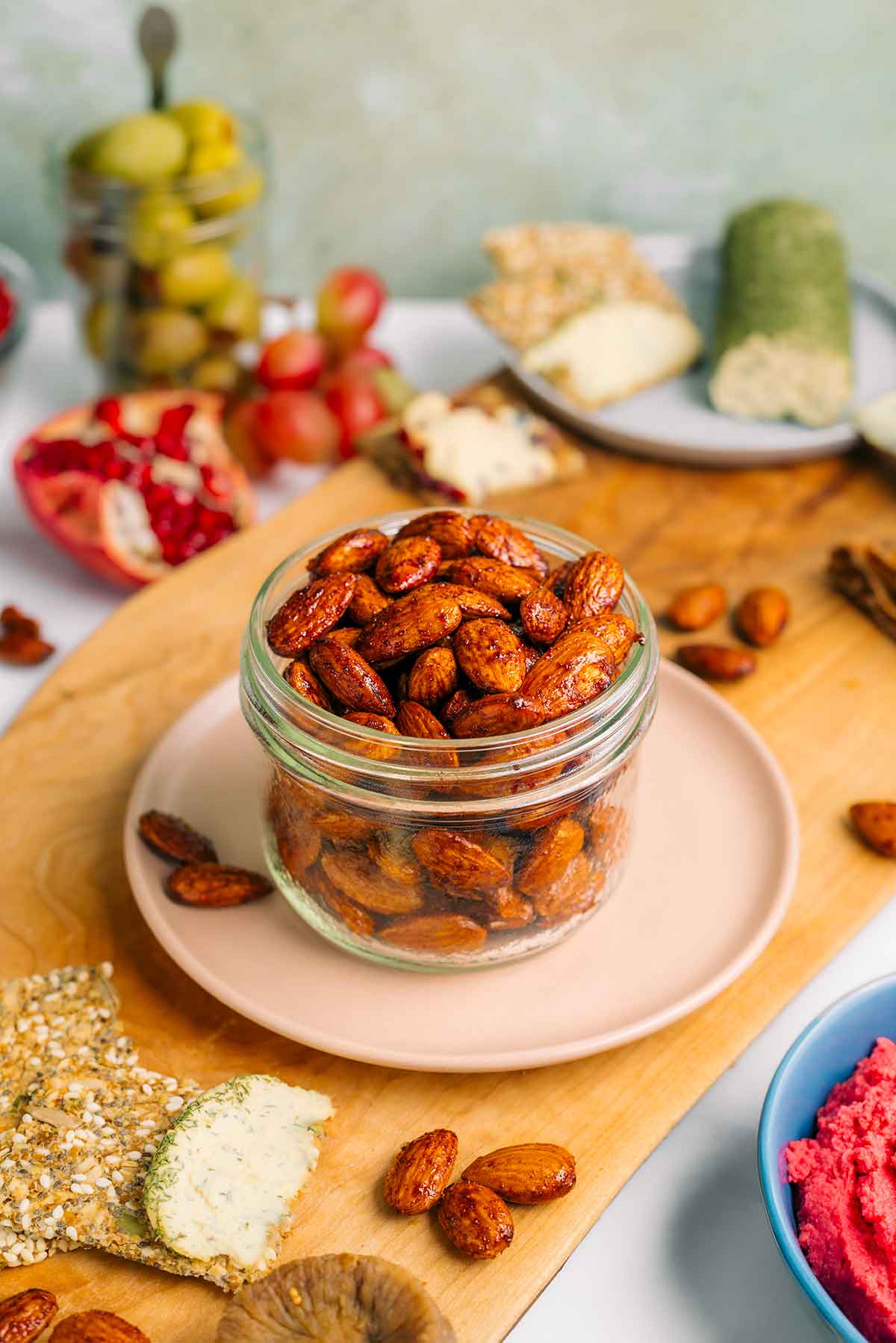 Side view of smoky maple almonds filling a small glass jar. The golden roasted almonds are shiny with a maple coating and speckled with a smoky flavouring. Charcuterie board items surround these spiced nuts.