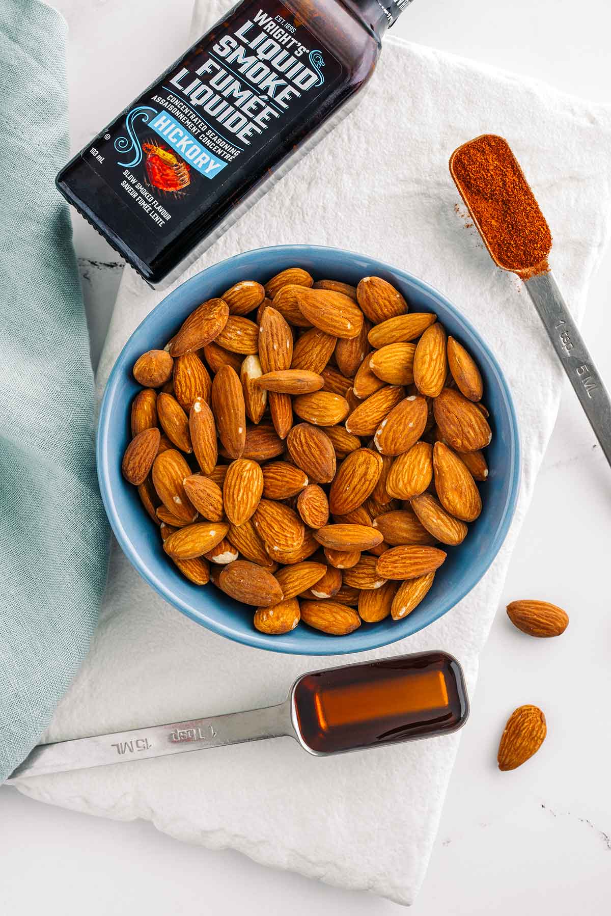 Top down view of ingredients. Raw almonds fill a bowl and are surrounded by maple syrup, smoked paprika, and a small bottle of Wright's liquid smoke.