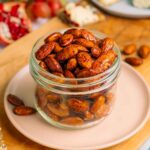 Side view of smoky maple almonds filling a small glass jar. The golden roasted almonds are shiny with a maple coating and speckled with a smoky flavouring.