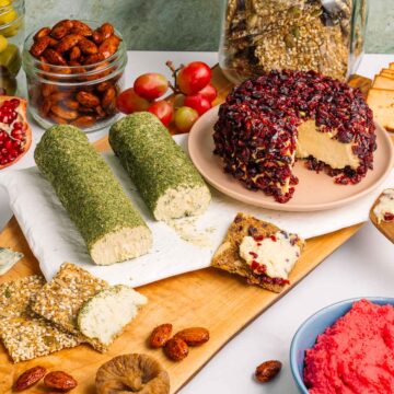 Side view of a full vegan charcuterie board. A cranberry crusted vegan cheese ball rests beside two garlic and dill vegan cheese logs. Homemade seed crackers are spread with the soft cheese. Nuts fruits, and a pink beet hummus surround.