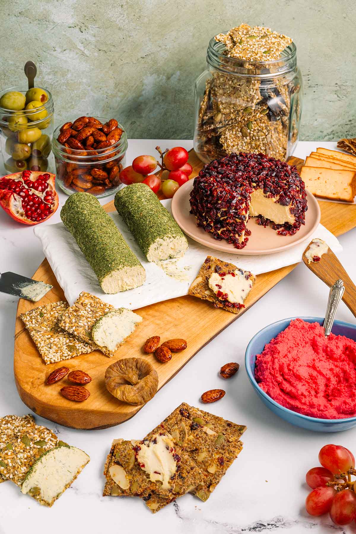 Side view of a full vegan charcuterie board. Two flavours of homemade vegan cheese are surrounded by homemade seed crackers, nuts, fruits, smoked tofu, and beet hummus.