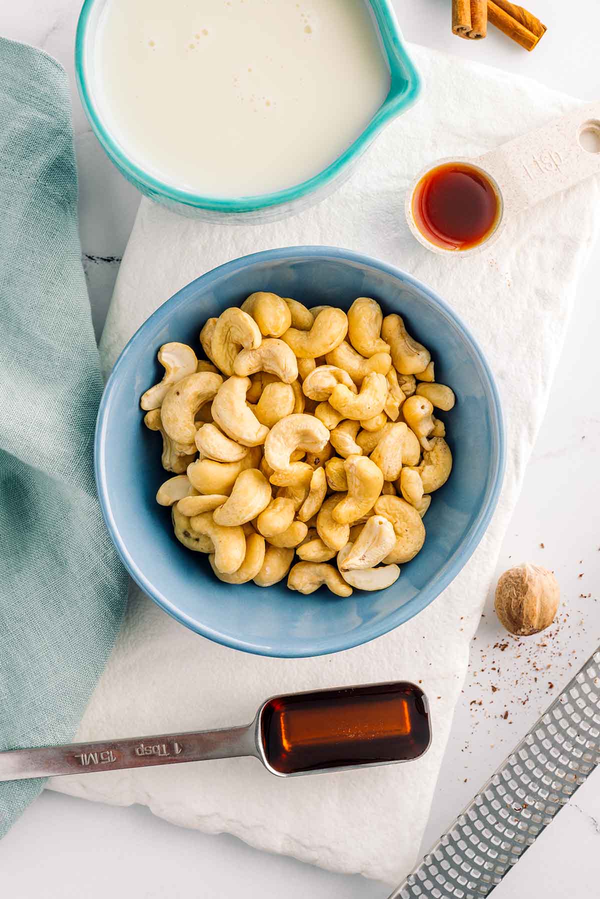 Top down view of ingredients. Raw cashews fill a small bowl with plant milk, maple syrup, nutmeg, and vanilla extract surrounding.