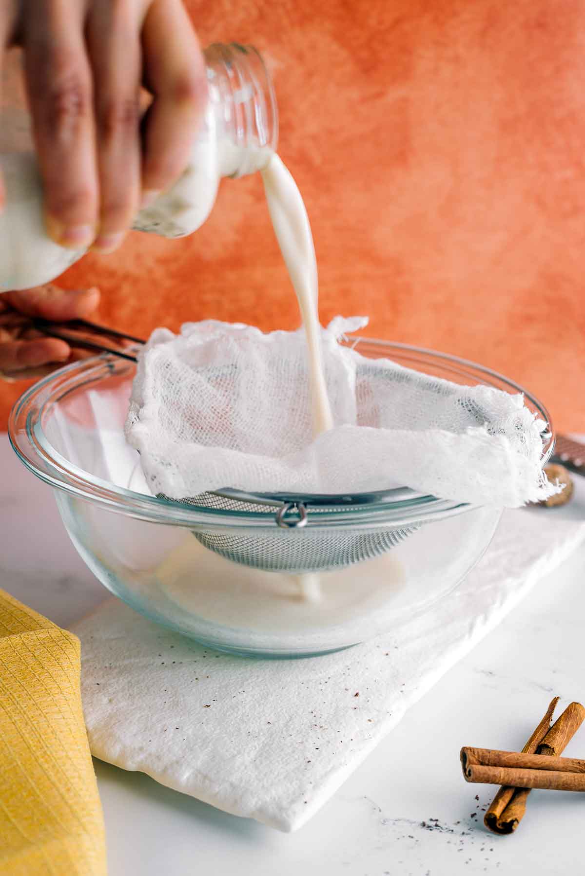 Side view of vegan cashew eggnog being poured through a fine mesh sieve lined with cheesecloth. The creamy eggnog flows through the lined sieve into a large bowl.