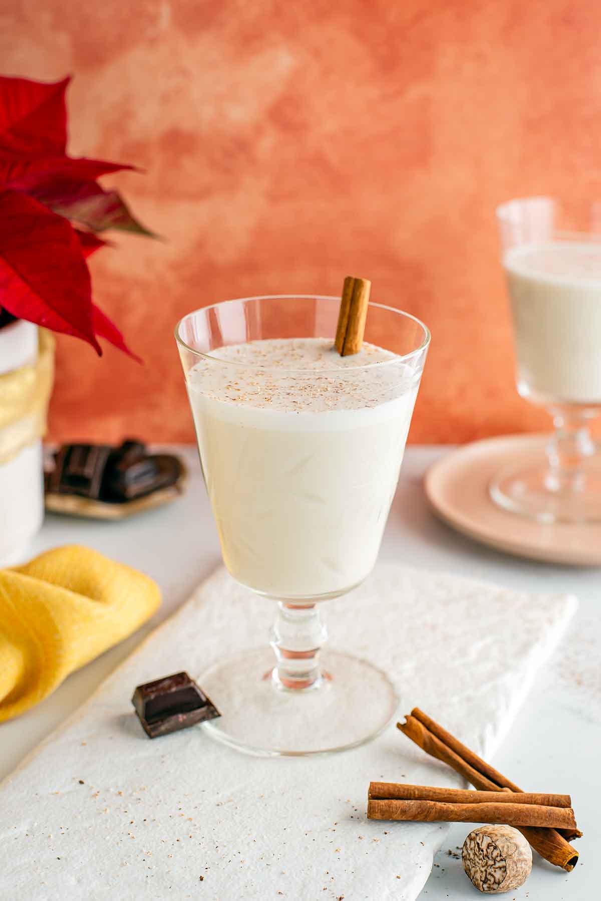 A beautiful glass is filled with creamy vegan eggnog garnished with fresh nutmeg and a cinnamon stick. Another glass is in the background and a square of chocolate rests against the bottom of the glass.