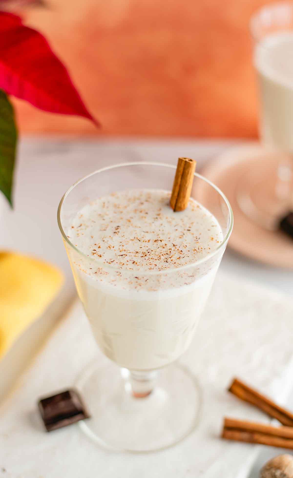 Top down view of creamy vegan eggnog in a beautiful glass. The eggnog is garnished with freshly grated nutmeg and a cinnamon sticks rests in the festive drink.