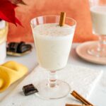 A beautiful glass is filled with creamy vegan eggnog garnished with fresh nutmeg and a cinnamon stick.