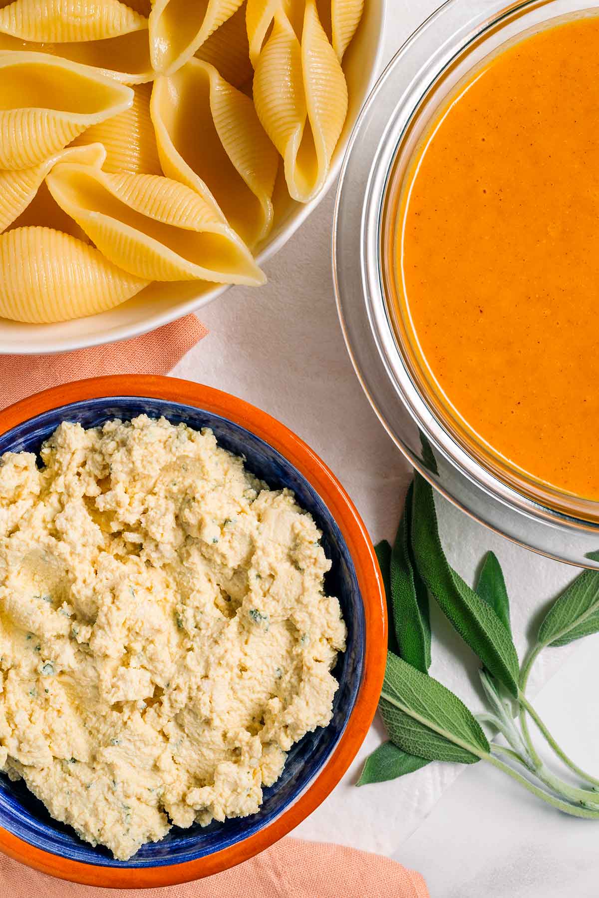 Top down view of ingredients. Tofu ricotta fills a small bowl beside a bowl of orange pumpkin sauce and a plate of cooked jumbo pasta shells. 