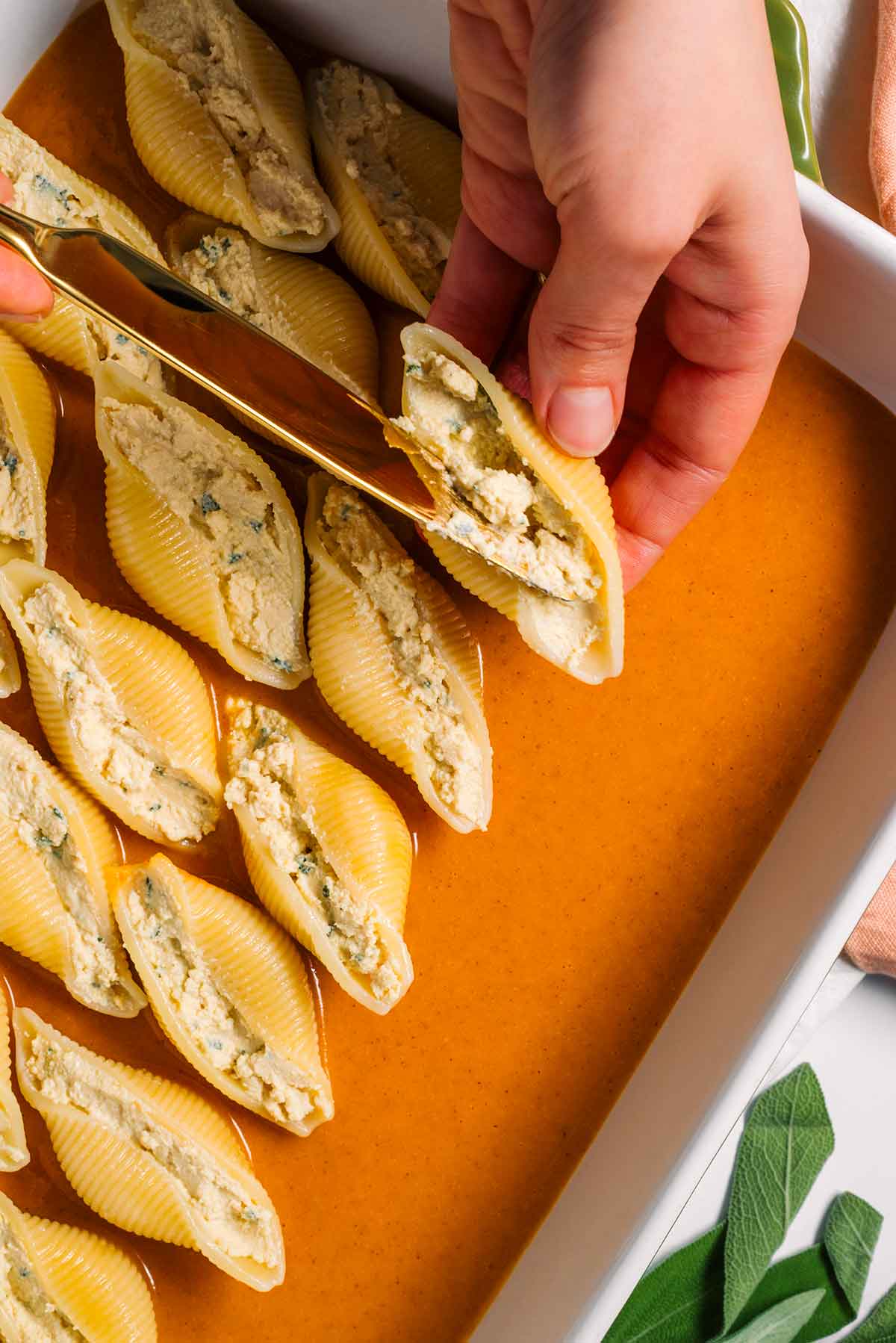 Top down view of hands filling a jumbo pasta shell with tofu ricotta and placing it in a casserole dish filled with a layer of pumpkin sauce.