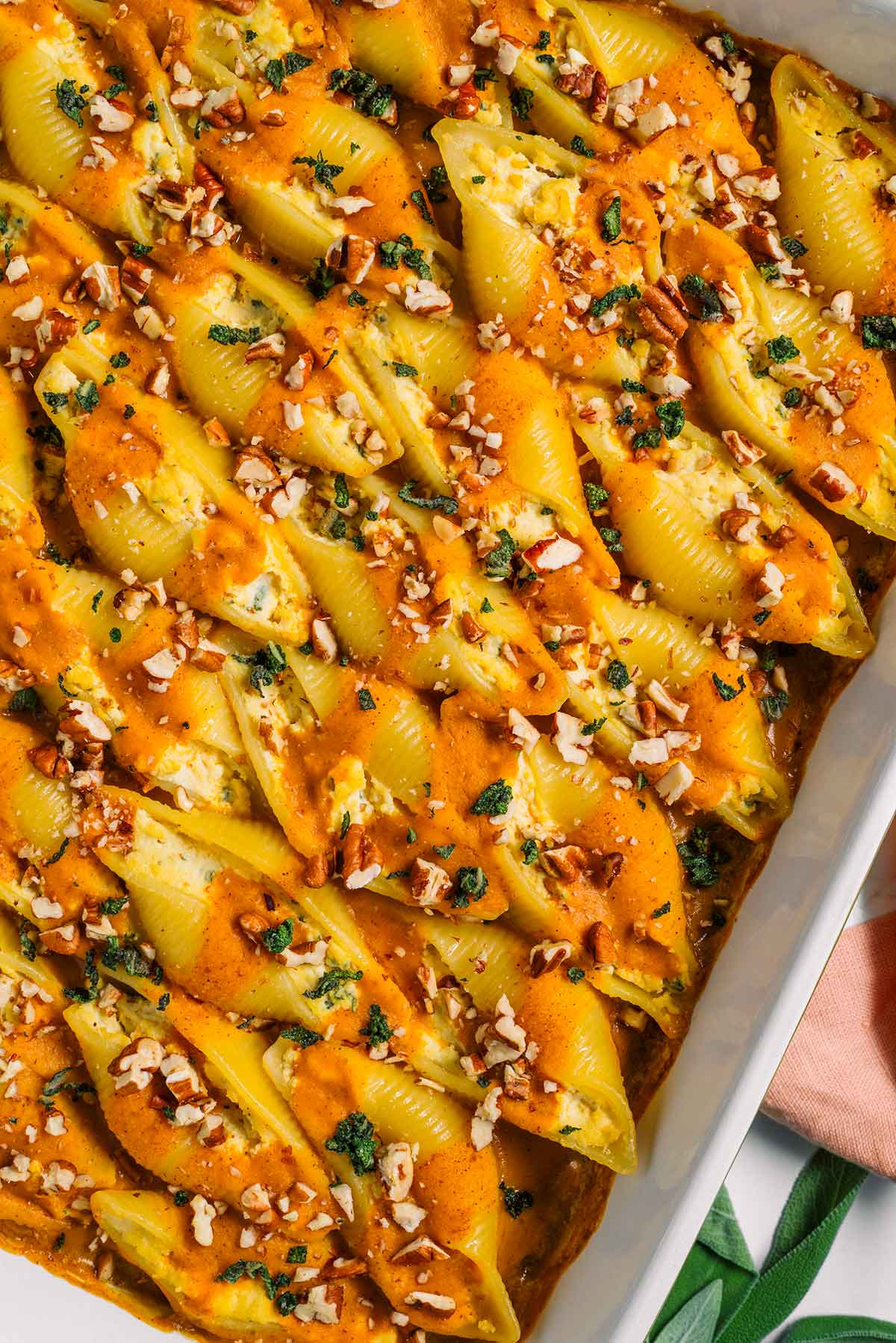 Top down view of vegan stuffed shells in a baking tray. A pumpkin sauce is delicately drizzled over the tofu ricotta stuffed shells and they are garnished with crispy sage and chopped toasted pecans.