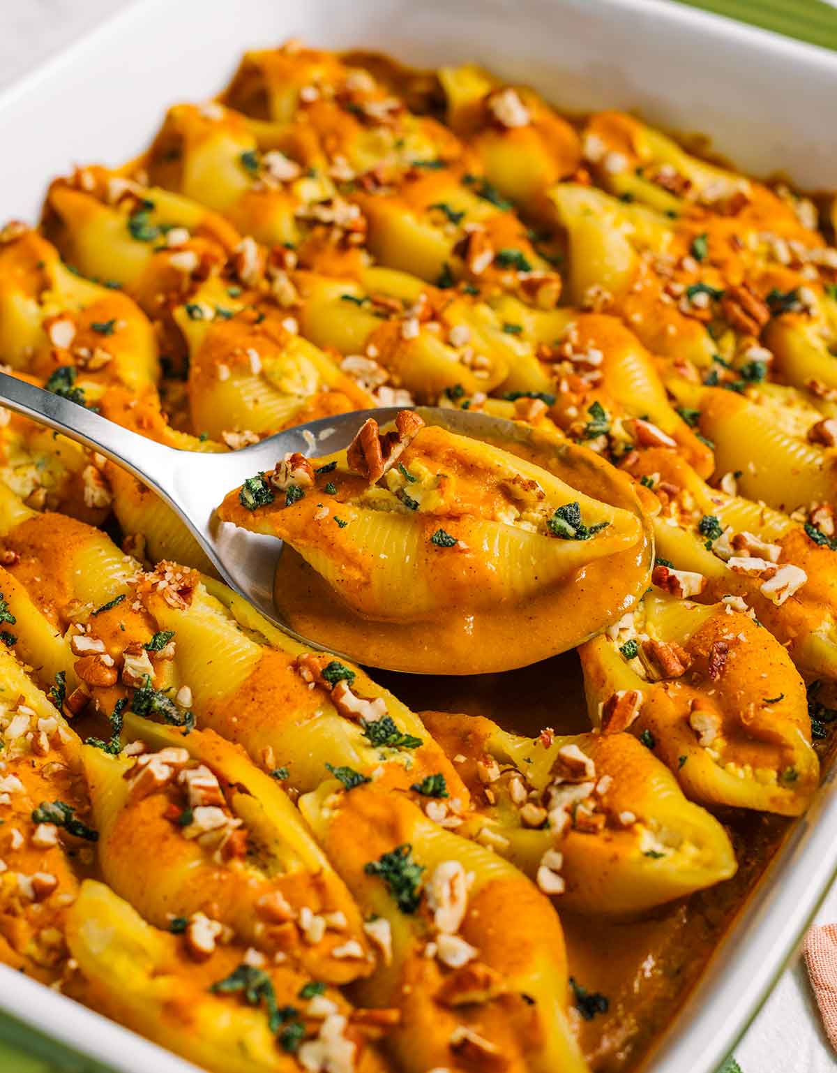 Side view of a spoon lifting a jumbo stuffed shell from a casserole dish. A pumpkin sauce fills the spoon and the ricotta stuffed shell delicately sits on top of the sauce.