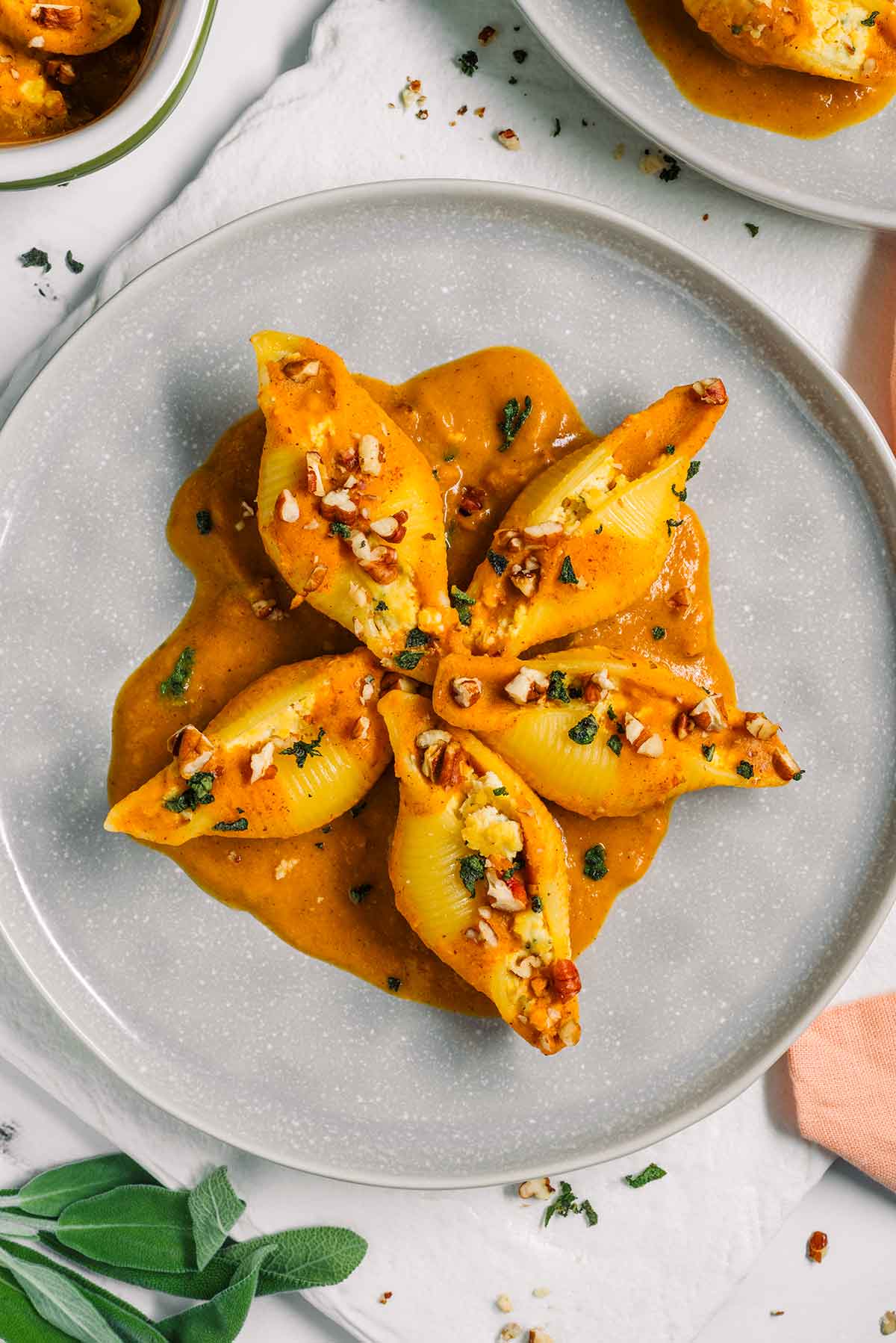 Top down view of a plate of 5 vegan stuffed shells arranged in a star patters. Pumpkin sauce is layered underneath the beautiful shells garnished with fresh sage and toasted pecans.