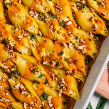 Top down view of vegan stuffed shells beautifully lined up in a baking tray. A pumpkin sauce is delicately drizzled over the tofu ricotta stuffed shells and they are garnished with crispy sage and chopped toasted pecans.