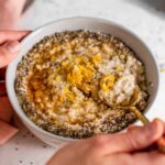 Closeup side view of a hand scooping a spoonful of lemon chia oatmeal topped with lemon zest, chia seeds, and maple syrup.