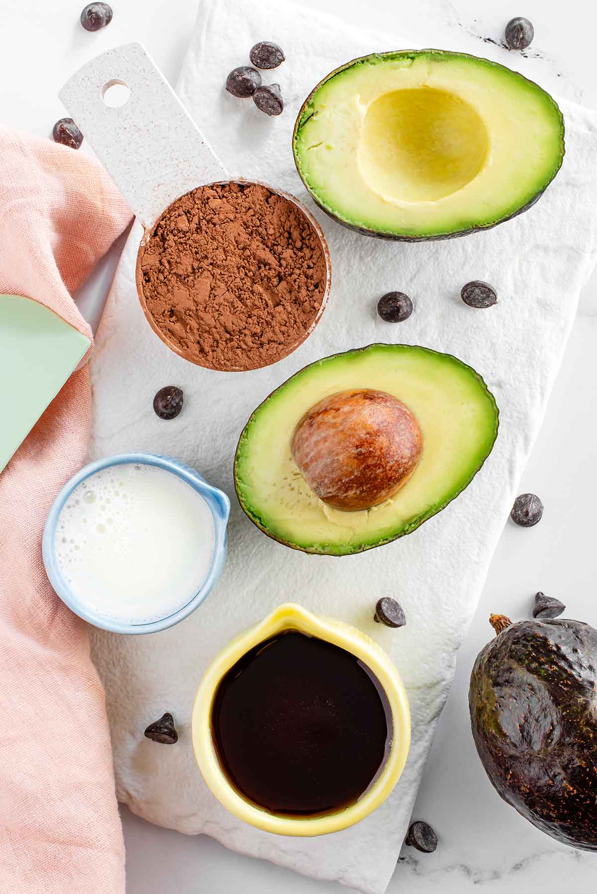 Top down view of ingredients for chocolate avocado pudding. An avocado is sliced in half and rests beside cocoa powder, plant milk, maple syrup, and scattered chocolate chips.