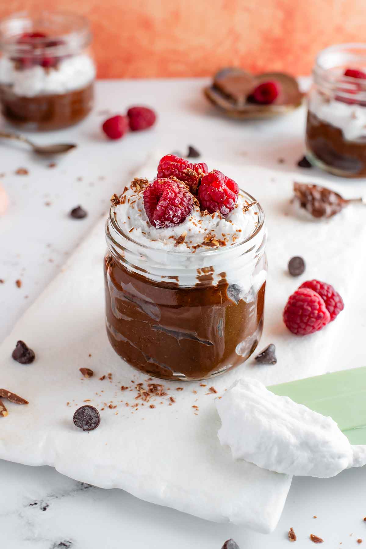 Side view of a thick and creamy chocolate avocado pudding in a small glass jar on top of a white tray.. The pudding is topped with fluffy coconut whipped cream, raspberries, and shaved chocolate. Other pudding cups can be seen in the background.