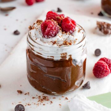 Side view of a thick and creamy chocolate avocado pudding in a small glass jar. The pudding is topped with fluffy coconut whipped cream, raspberries, and shaved chocolate.