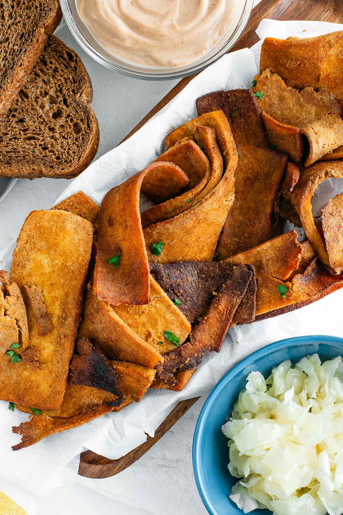 Top down view of ingredients for a vegan reuben. Thin pieces of tofu shaved meat are swirled on a tray surrounded by sauerkraut, Russian dressing, and dark rye bread.