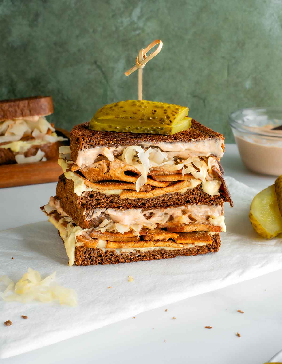 Side view of a sliced vegan reuben. The two halves are stacked on top of one another and a pickle is secured to the top of the gooey and loaded sandwich with a skewer.