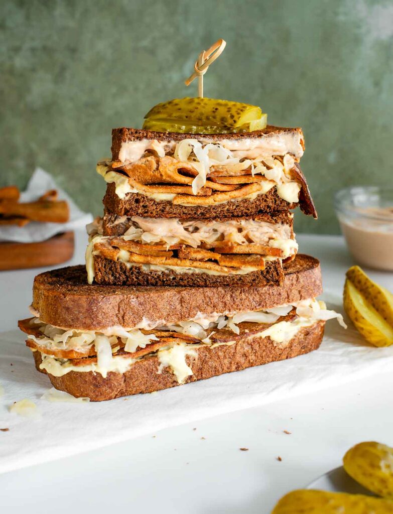Two vegan reuben sandwiches are stacked on a white tray. Gooey cheese oozes from the sides and strips of sauerkraut can be seen on top of a tofu meat. Creamy Russian dressing peeks out from under dark rye bread and pickles are skewered on top of the sandwich.