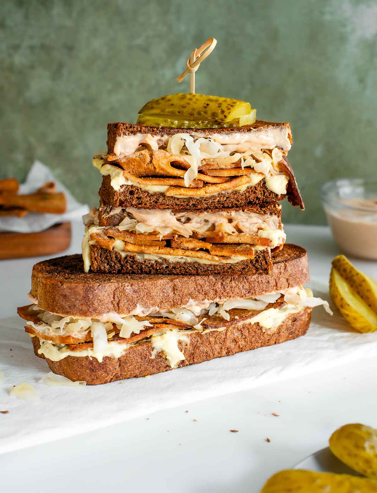 Two vegan reuben sandwiches are stacked on a white tray. Gooey cheese oozes from the sides and strips of sauerkraut can be seen on top of a tofu meat. Creamy Russian dressing peeks out from under dark rye bread and pickles are skewered on top of the sandwich. 