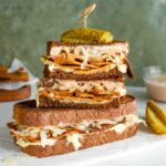 Two vegan reuben sandwiches are stacked on top of each other. Gooey cheese oozes from the sides and strips of sauerkraut can be seen on top of a tofu meat. Creamy Russian dressing peeks out from under dark rye bread and pickles are skewered on top of the sandwich.