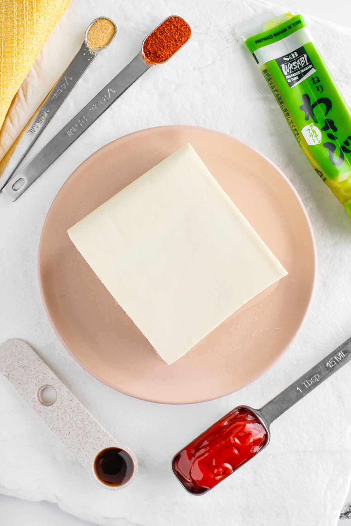 Top down view of ingredients for vegan Russian dressing. A block of soft tofu rests on a plate surrounded by ketchup, worcestershire sauce, onion powder, smoked paprika, and wasabi.