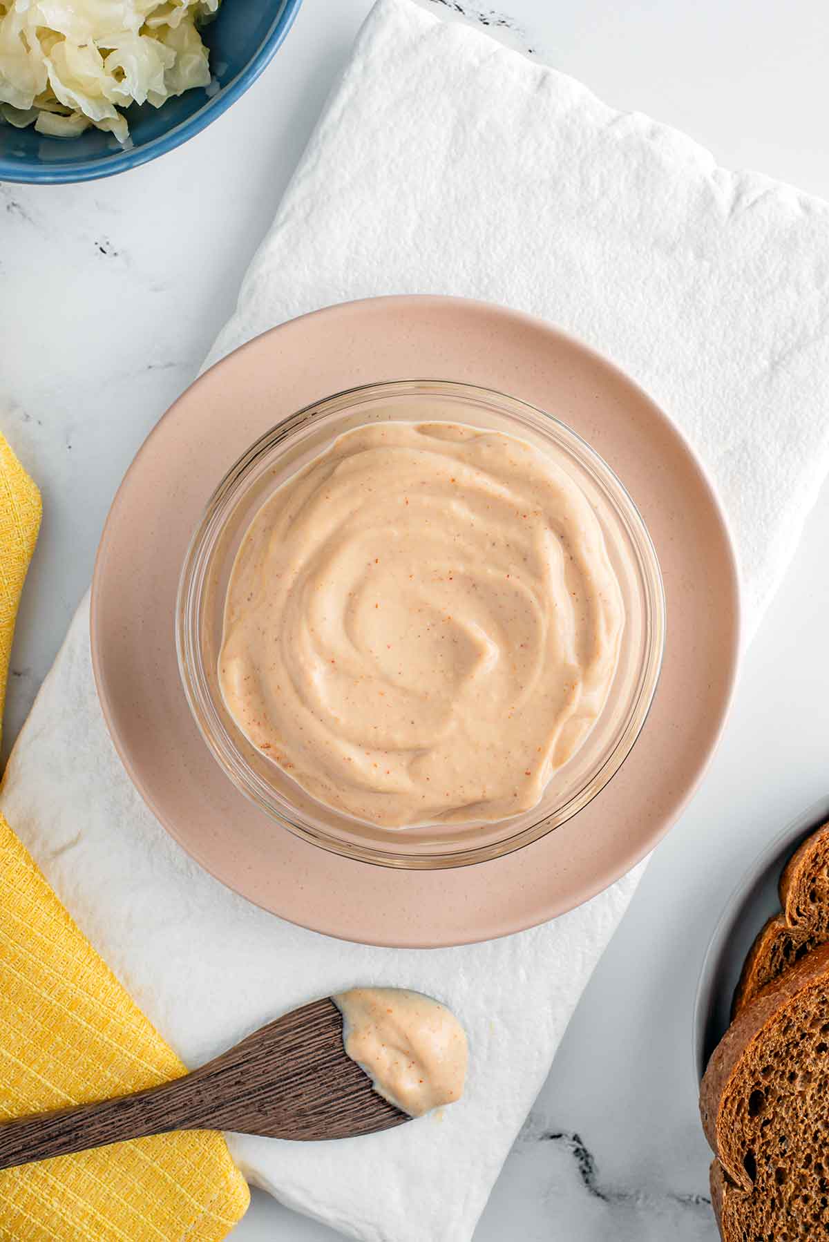 Top down view of thick and creamy vegan Russian dressing in a small bowl. The dressing has a light pink hue and is speckled with seasonings. Ingredients for a reuben sandwich are scattered around the bowl of dressing.