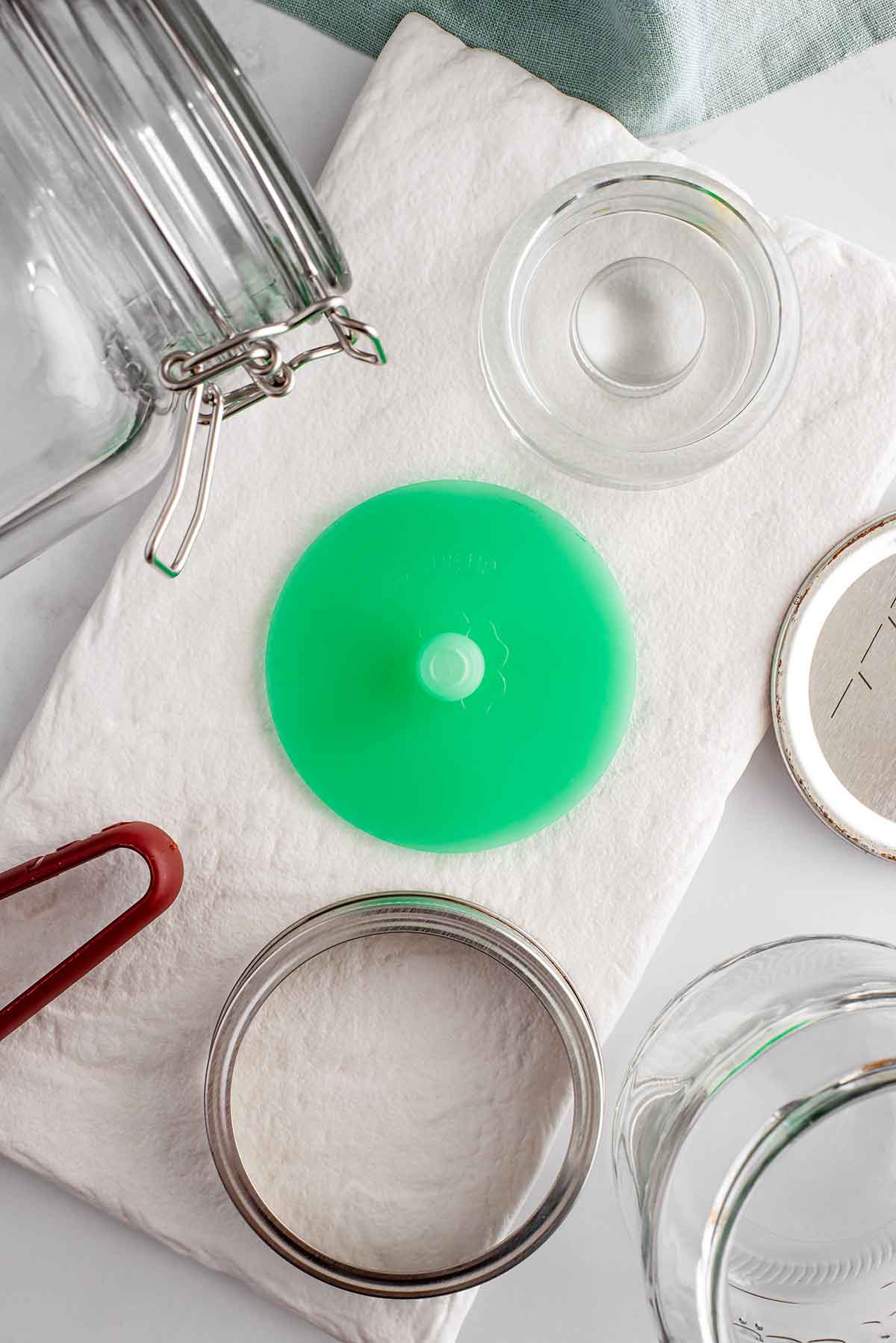 Top down view of fermentation tools. A pickle pipe fermenting lid, a glass fermentation weight, a wide mouth glass jar, and a clamp lid glass jar are gathered on a white tray.