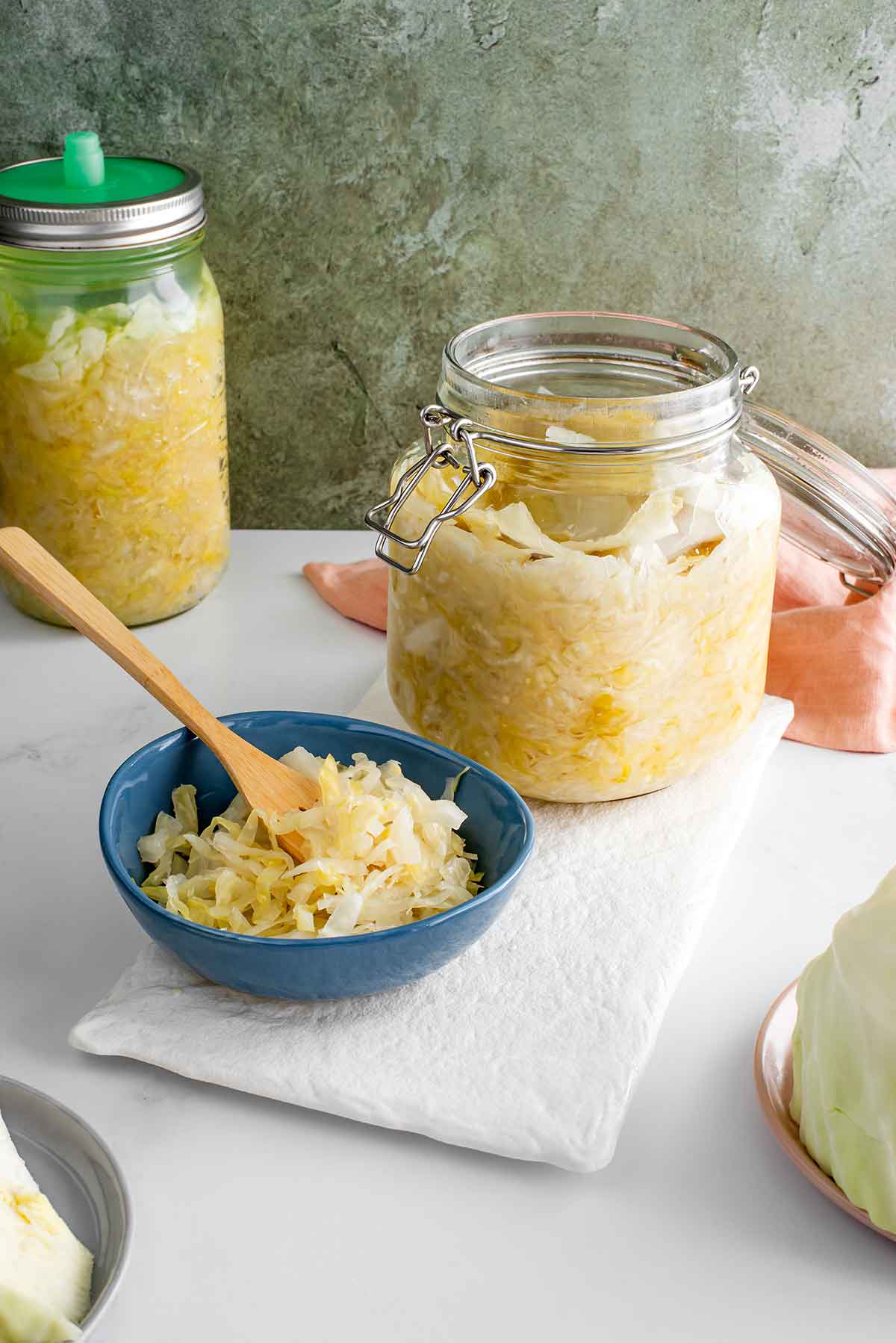 A jar of homemade sauerkraut rests open on a white tray. A small bowl of sauerkraut with a wooden fork resting in the fermented cabbage stands in front of the jar. In the background is a jar of sauerkraut partway through the fermentation process with a pickle pipe lid.