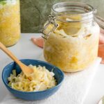 A jar of homemade sauerkraut rests open on a white tray. A small bowl of sauerkraut with a wooden fork resting in the fermented cabbage stands in front of the jar.