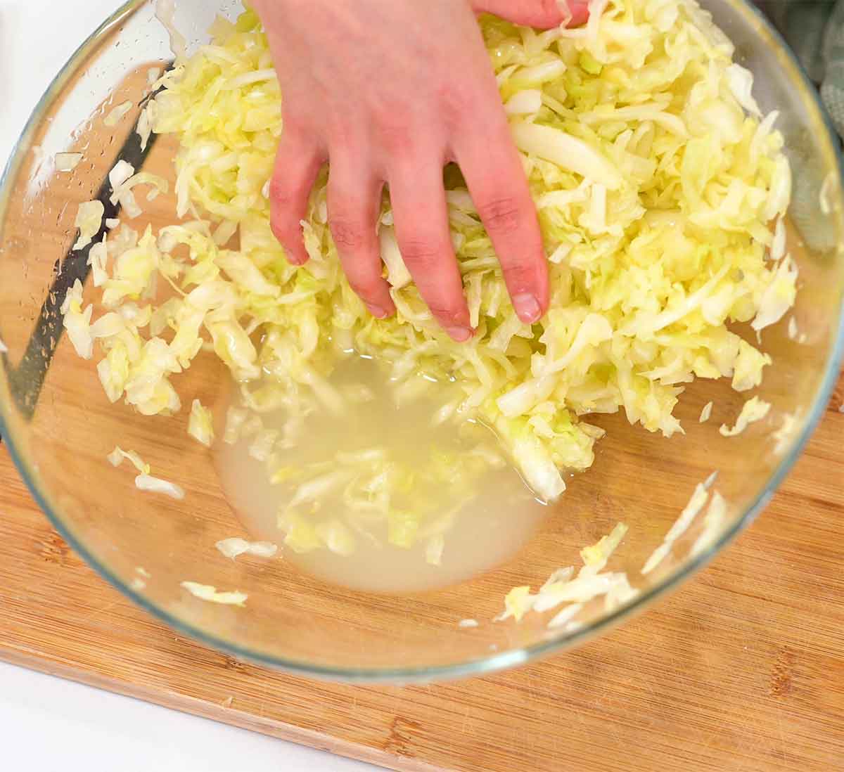 Top down view of a hand pulling massaged shredded cabbage to the side revealing liquid gathered at the bottom of a bowl.