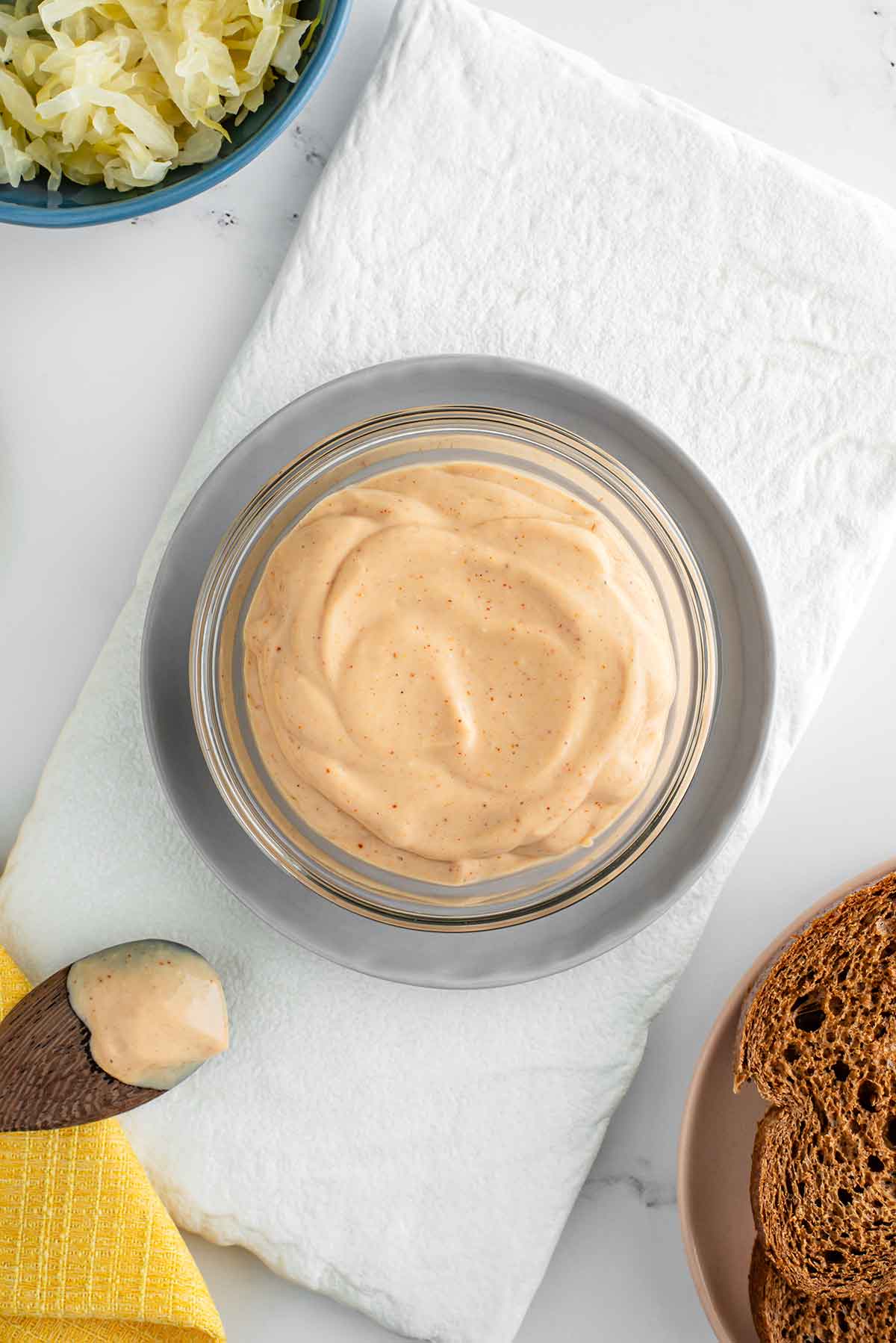 Top down view of a bowl of thick and creamy vegan Russian dressing. The dressing is tinged pink and is speckled with spices. Ingredients for a vegan Reuben are placed around the bowl. 