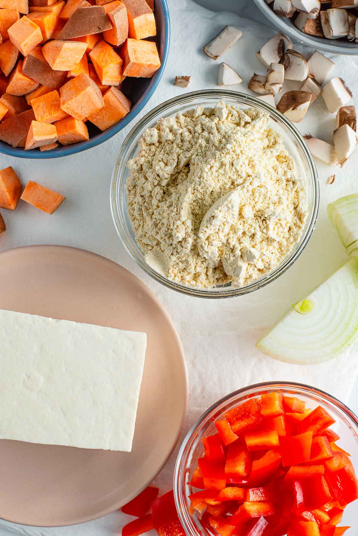 Top down view of ingredients on a white tray. Tofu and chickpea flour are surrounded by small diced sweet potato, bell pepper, mushrooms, and onion.