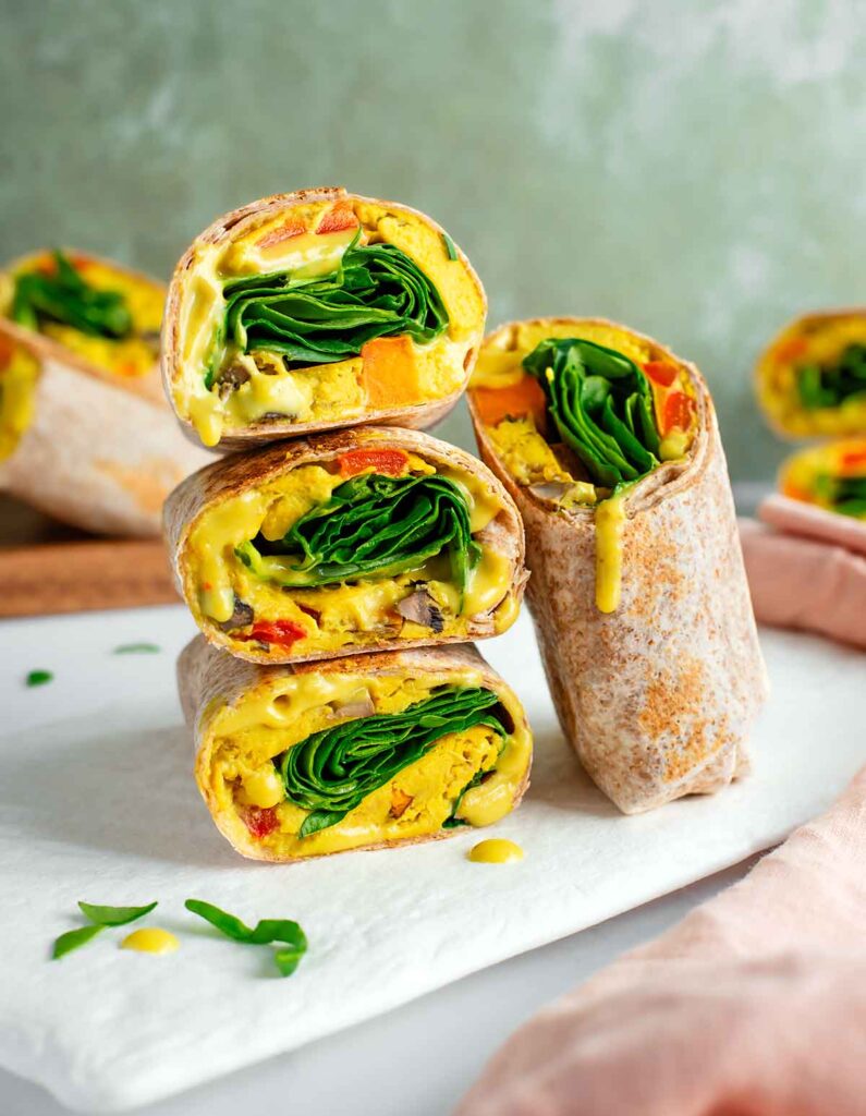 Side view of stacked vegan breakfast burritos. They are filled with a vegan egg and vegetable patty, fresh spinach, and a runny vegan queso. More burritos wrapped in whole wheat tortilla fill the background.