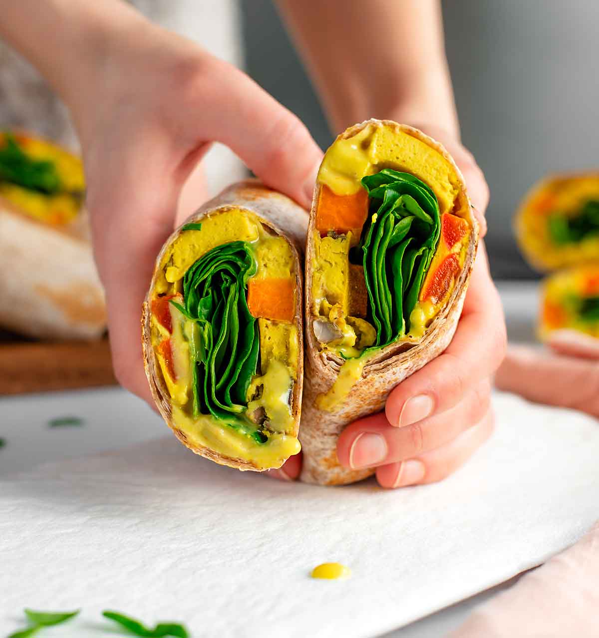 Side view of hands holding a sliced vegan breakfast burrito revealing the fillings. A vegan egg and vegetable patty fills the whole wheat tortilla along with a runny vegan queso and fresh spinach.