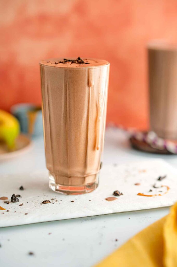 Side view of a glass filled to the brim with a thick vegan chocolate milkshake. Thick drips slide down the top of the glass. Chocolate shavings garnish the shake and are sprinkled on the white tray beneath. Another milkshake stands in the background.