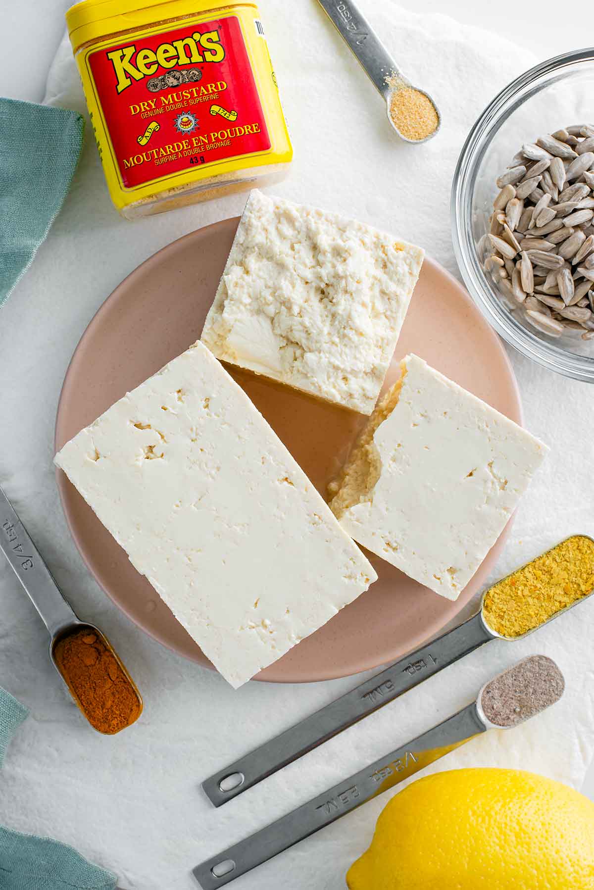 Top down view of medium firm tofu on a small plate. The tofu is loosely broken apart to show its soft texture. Surrounding the tofu is a bowl of sunflower seeds, a package of mustard powder, a lemon, and other seasonings in measuring spoons.