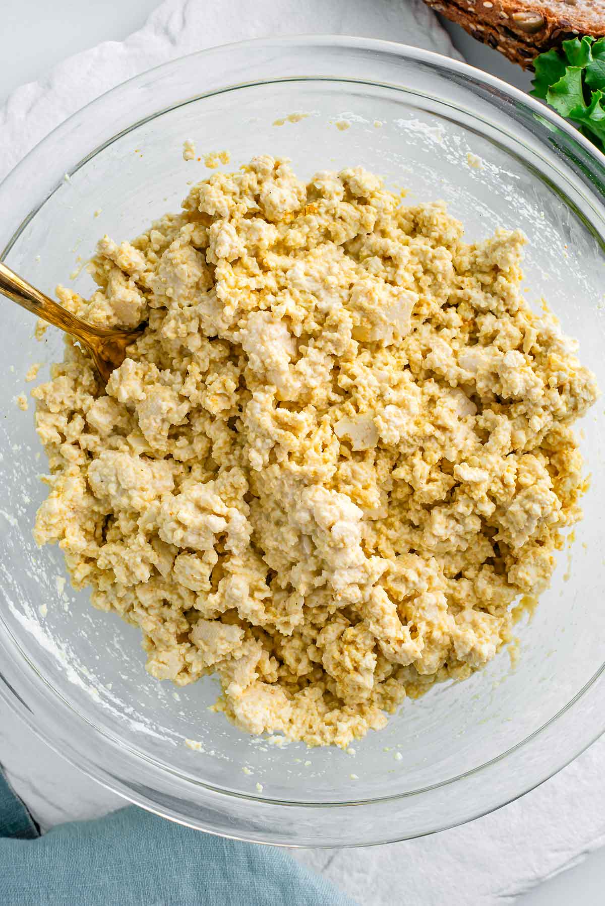 Top down view of creamy vegan tofu egg salad in a glass mixing bowl. A fork rests in the mashed tofu which is a light yellow colour. Some slightly larger pieces of tofu are more intact and more white in colour. 