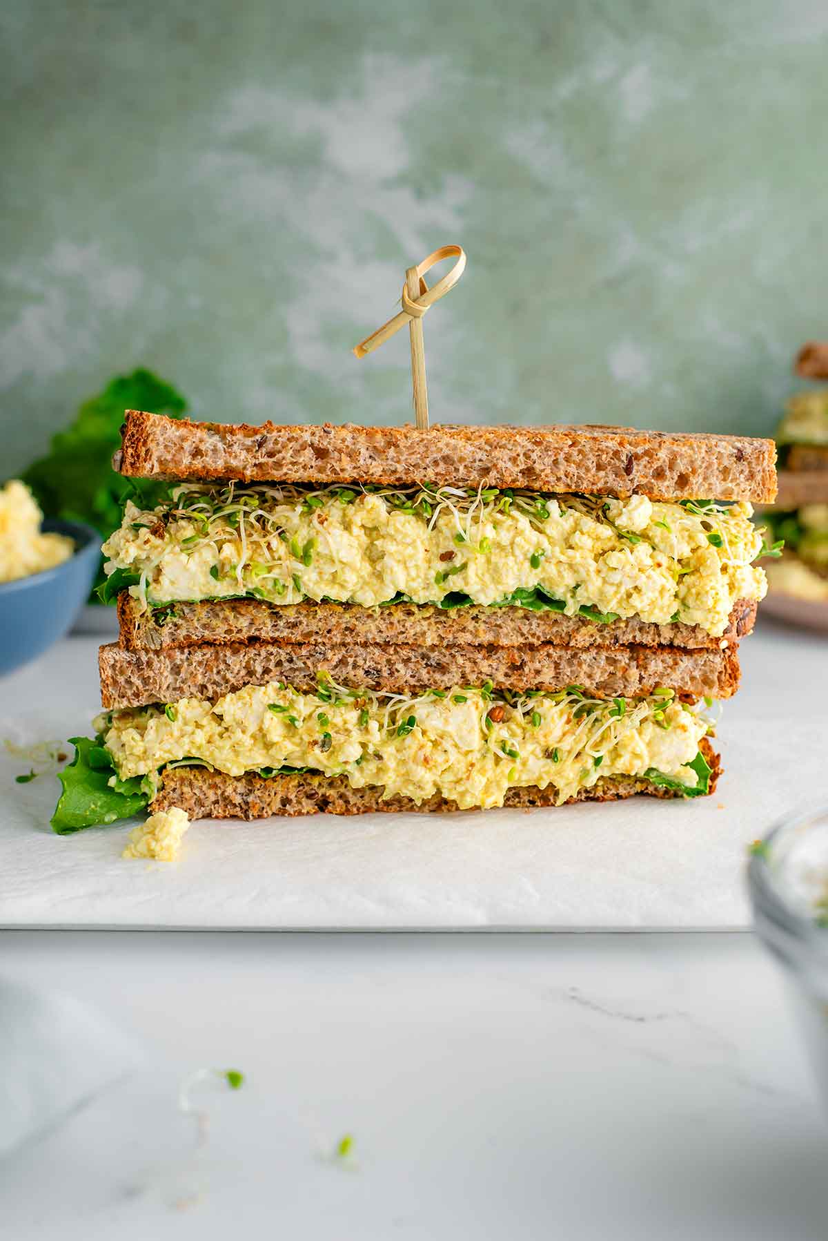 Side view of a vegan tofu egg salad sandwich. The tofu is a light yellow colour and a soft and mashed but not mushy texture. The sandwich bread is lined with green leaf lettuce and sprouts. A bamboo pick protrudes from the top of the sandwich and extra ingredients fill the background.
