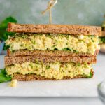 Side view of a vegan tofu egg salad sandwich. The tofu is a light yellow colour and a soft and mashed but not mushy texture. The sandwich bread is lined with green leaf lettuce and sprouts.