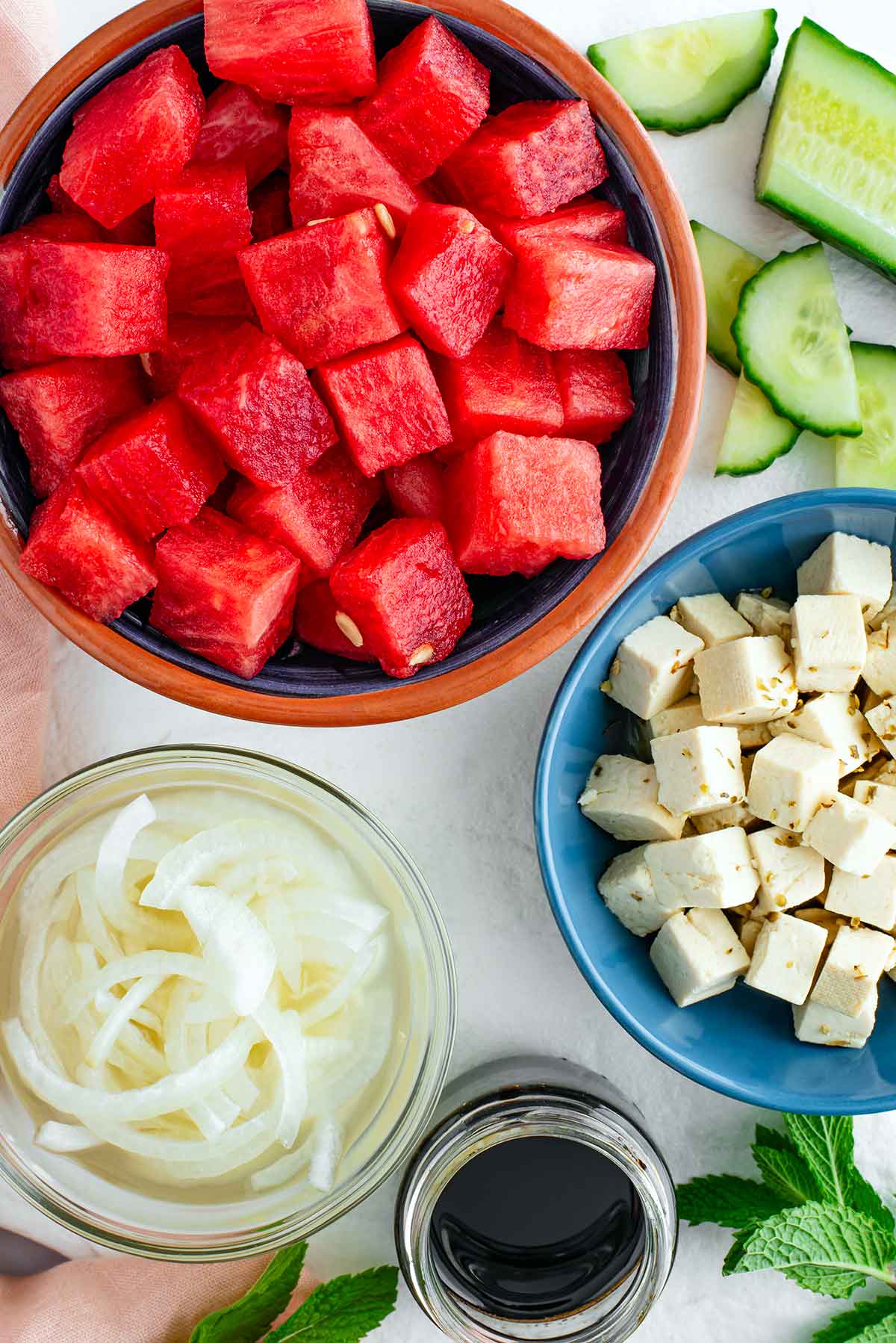 Top down view of cubed watermelon, cubed tofu feta, pickled white onion, sliced cucumber, fresh mint, and a small jar of balsamic maple reduction.