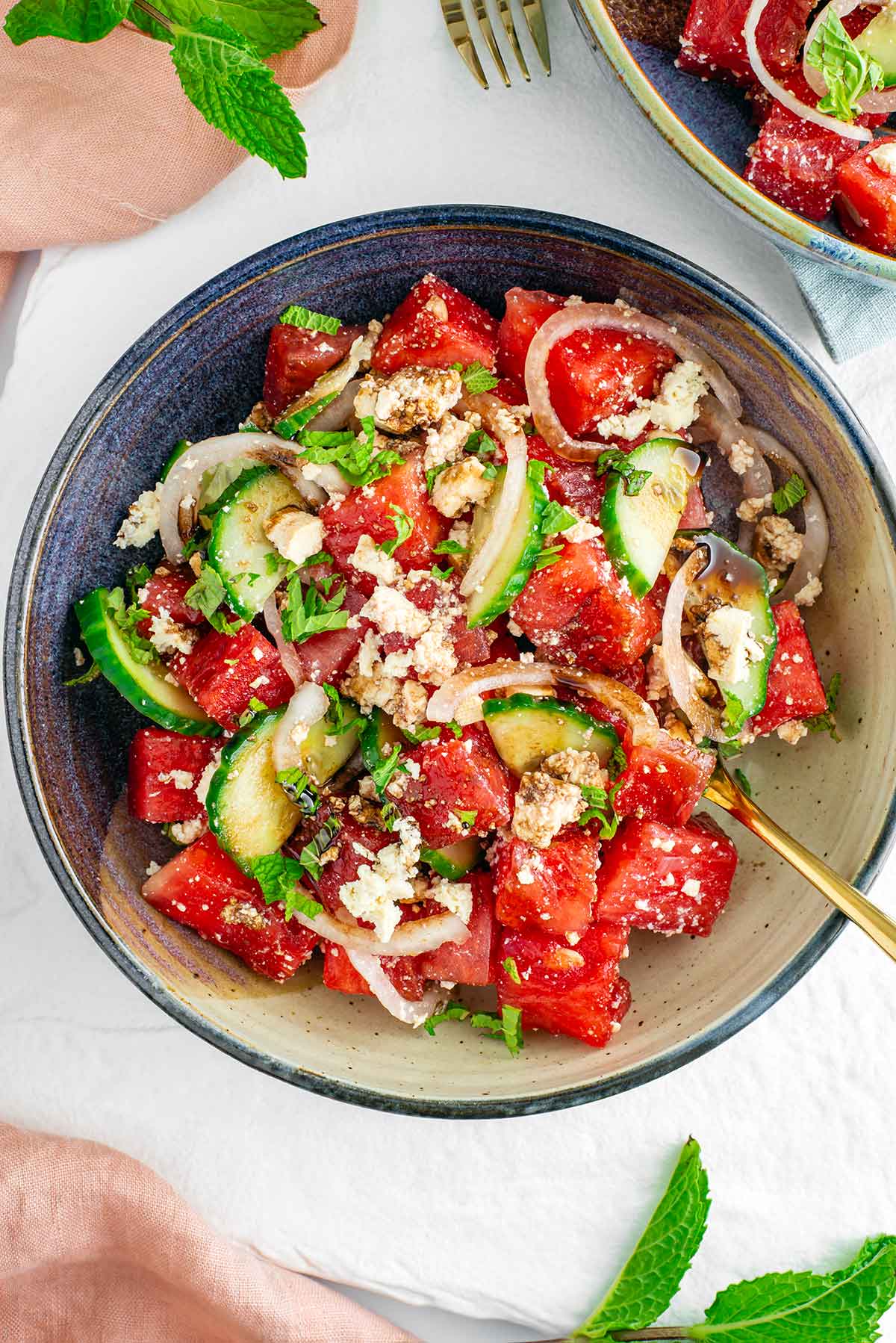 Top down view of watermelon salad with tofu feta filling a small bowl. Thinly sliced cucumber and pickled white onion add bulk and colour to the salad which is garnished with fresh mint and a balsamic reduction. Another bowl of salad is visible in the top corner of the photograph.