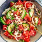 Top down view of watermelon salad with tofu feta filling a small bowl. Thinly sliced cucumber and pickled white onion add bulk and colour to the salad which is garnished with fresh mint and a balsamic reduction.
