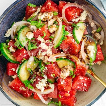 Top down view of watermelon salad with tofu feta filling a small bowl. Thinly sliced cucumber and pickled white onion add bulk and colour to the salad which is garnished with fresh mint and a balsamic reduction.