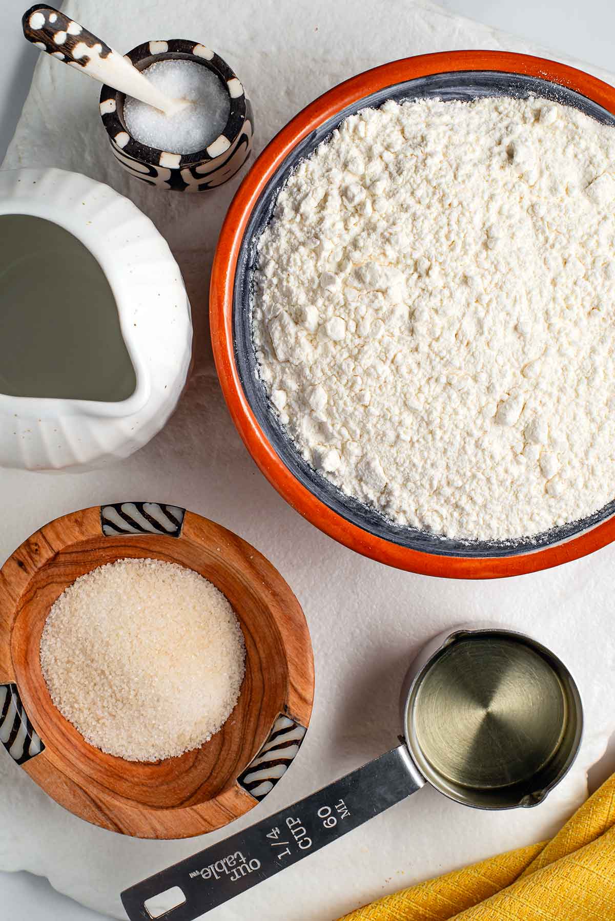 Top down view of ingredients on a white tray. All-purpose flour, water, sugar, salt, and vegetable oil. Some of the ingredients are in beautiful wooden dishes from Kenya.