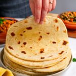 Side view of a hand lifting a soft and pliable chapati from a stack of Kenyan chapati. The chapati are nicely browned in spots and layered.
