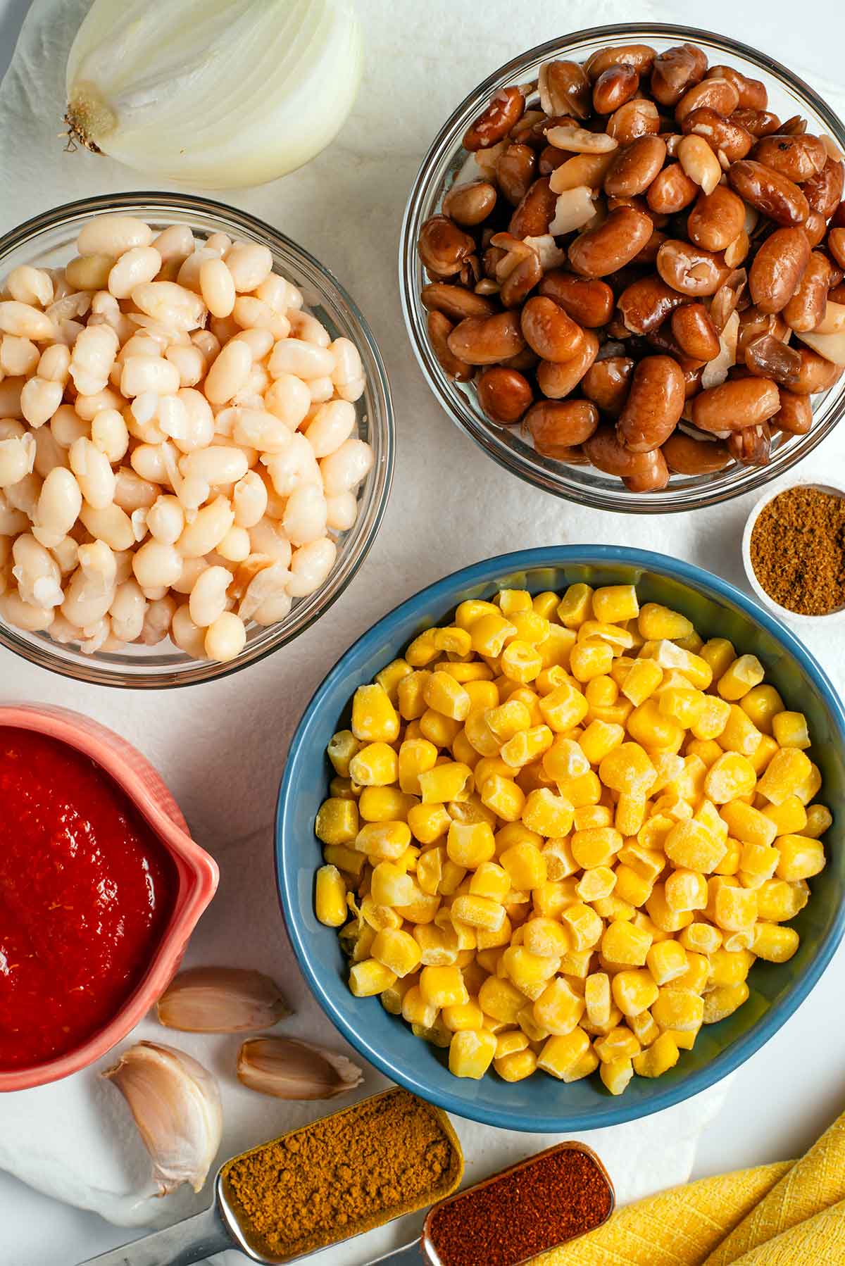 Top down view of ingredients on a white tray. Small bowls of corn, pinto beans, navy beans, and crushed tomatoes are gathered with onion, garlic, and small spoons of spices.